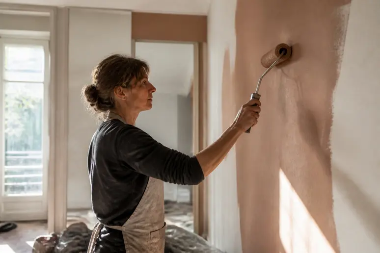 Une femme applique de la peinture terracotta satinée sur un mur de salon, lumière naturelle.