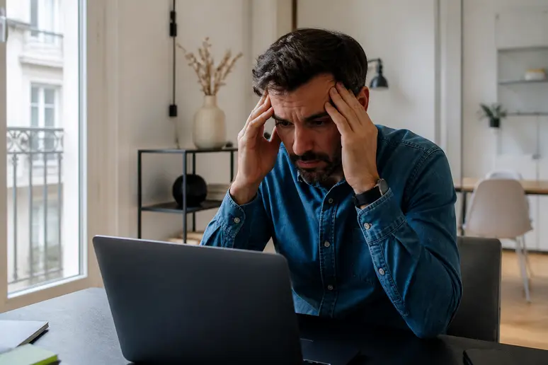Un homme d'une trentaine d'années froisse ses tempes à son bureau, dégageant une fatigue face à un angle de meuble.
