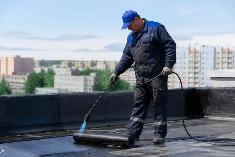 Artisan couvreur appliquant un rouleau de bitume sur un toit plat avec un chalumeau à gaz.